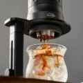 Schwarzes IKAPE Brühgerät auf einem Ständer mit dem Sieb unten, Espresso tropft in ein Glas mit Eis
