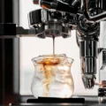 Dünner Espressostrahl aus dem Sieb in ein Glas mit Eis unter einer verchromten Espressomaschine