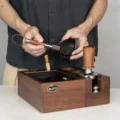 Person bürstet eine kleine Tasse über einer Organizer-Box aus Holz mit Tamper und anderen Werkzeugen aus
